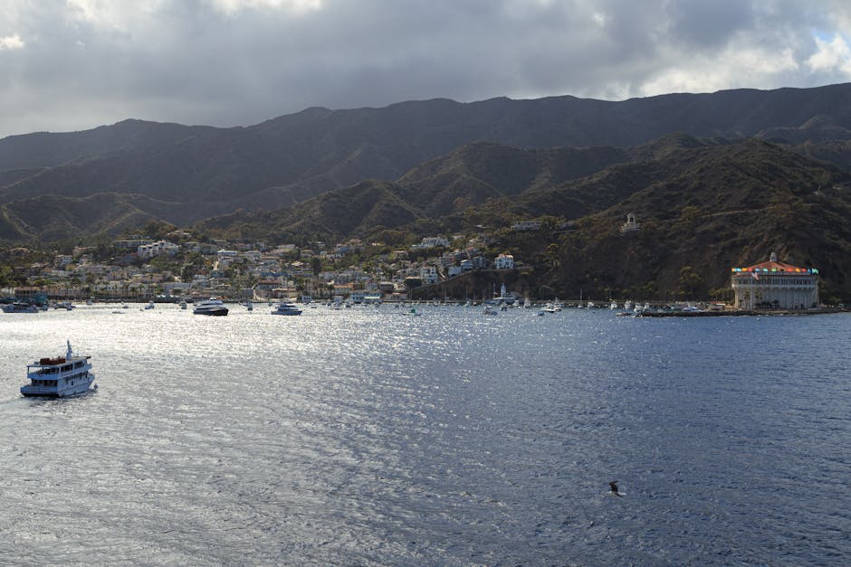 A serene harbor with boats, mountains, and a historic casino building on a cloudy day.