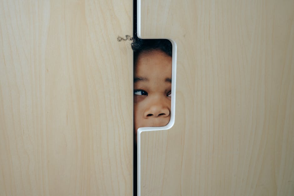 Anonymous funny little ethnic child hiding in wardrobe and peeping through hole while playing at home