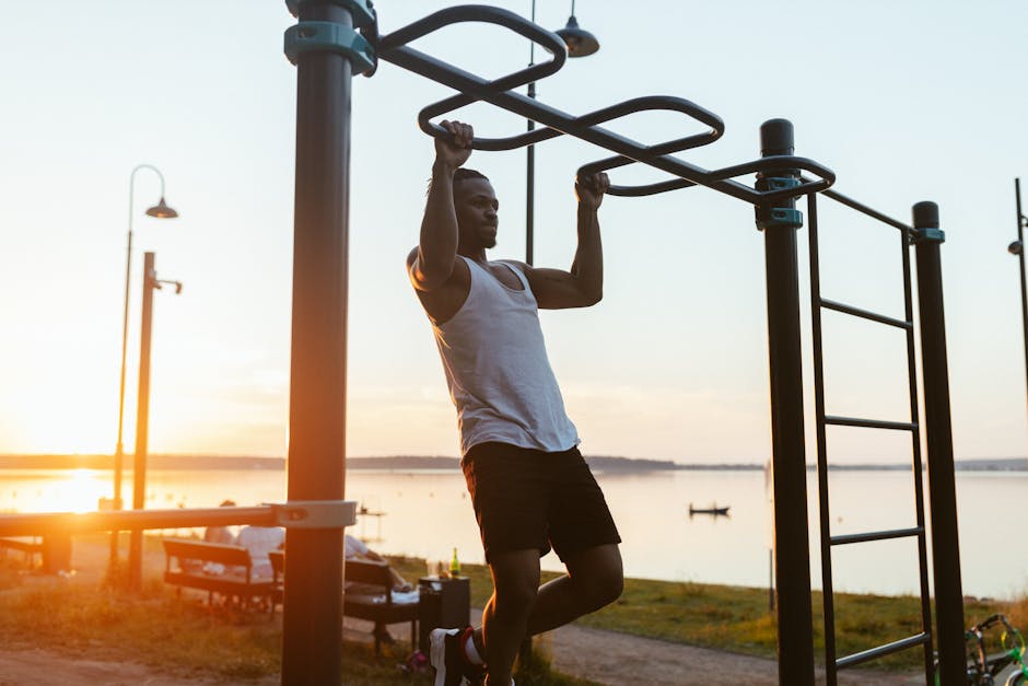 Man performing pull-ups on outdoor gym equipment by the lake during sunset.