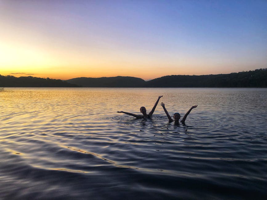 Two silhouetted people swimming in a serene lake at sunset, capturing a tranquil moment in nature.